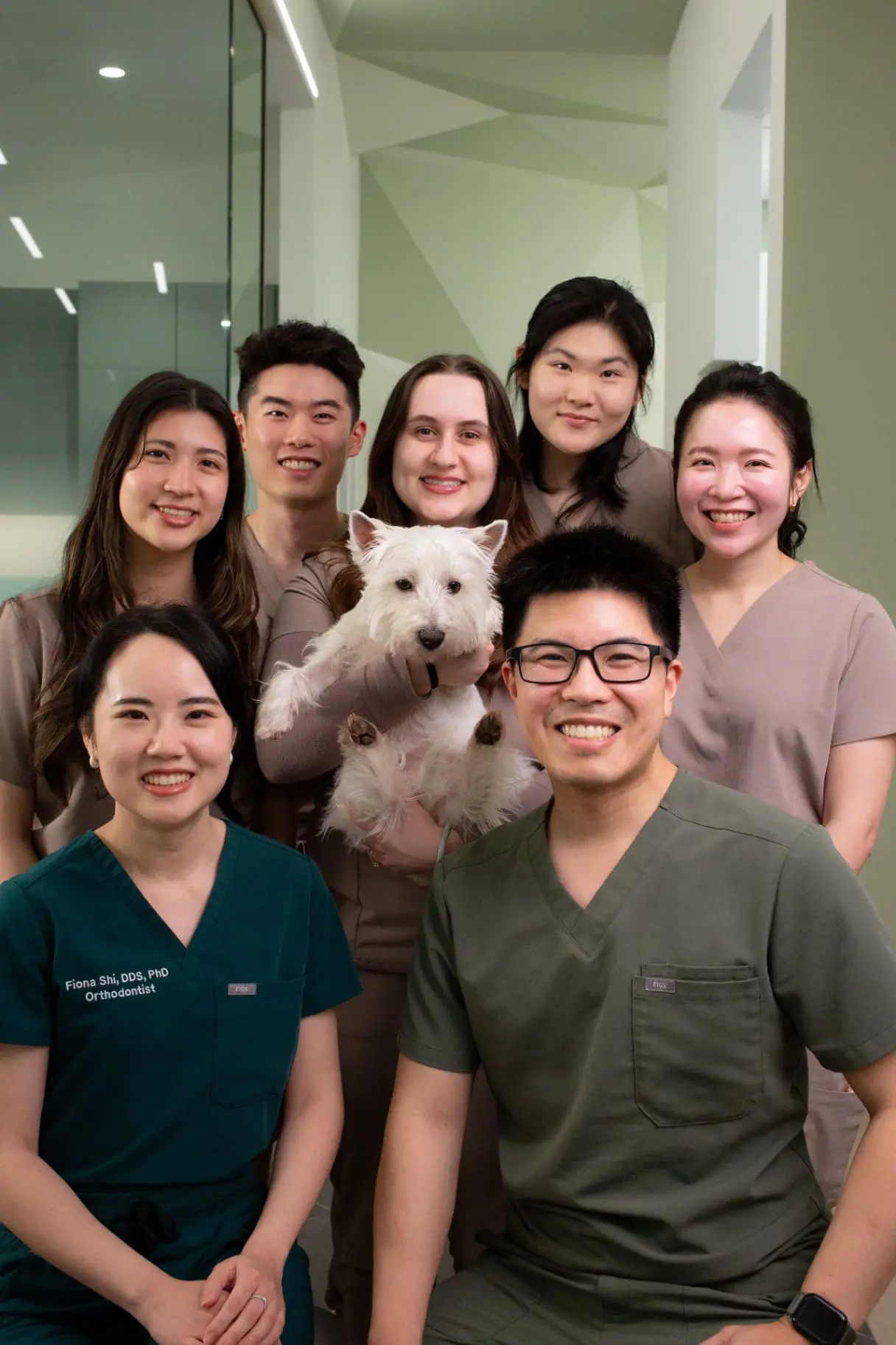 In Kirkland WA, the Emerald City Orthodontics team in scrubs smiles while posing with a small white dog in a bright hallway.