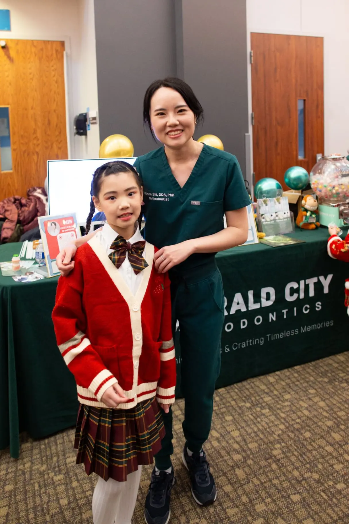 Dr. Jiayu (Fiona) Shi, orthodontist expert at Emerald City Orthodontics in Kirkland WA, poses with a young girl during a community event, standing in front of a decorated booth with balloons and informational displays.