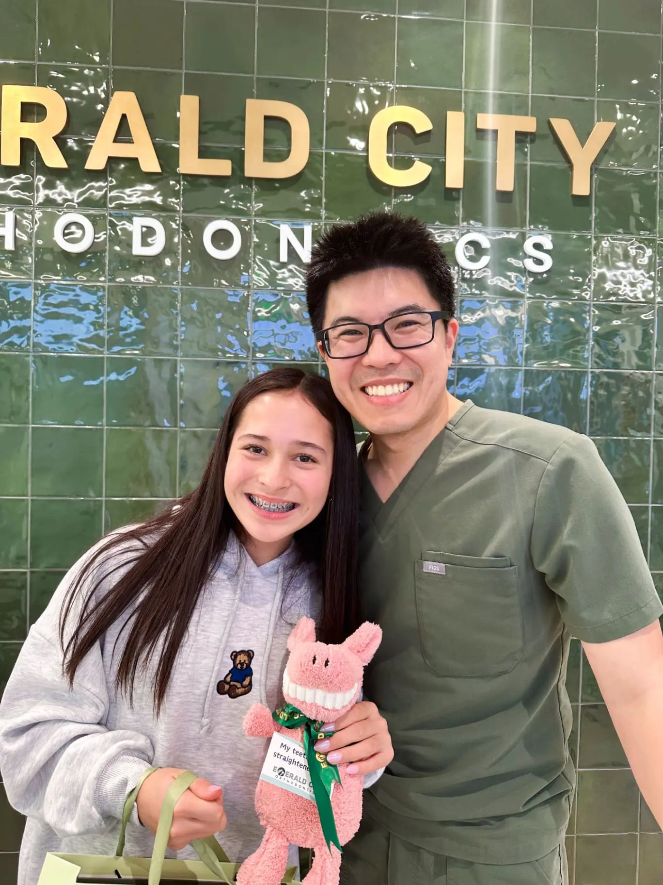 Dr. Hsin-Chuan (Dan) Pan, orthodontist expert at Emerald City Orthodontics in Kirkland WA, smiles with a happy patient holding a plush toy and gift bag in front of the clinic’s green tiled wall and logo.