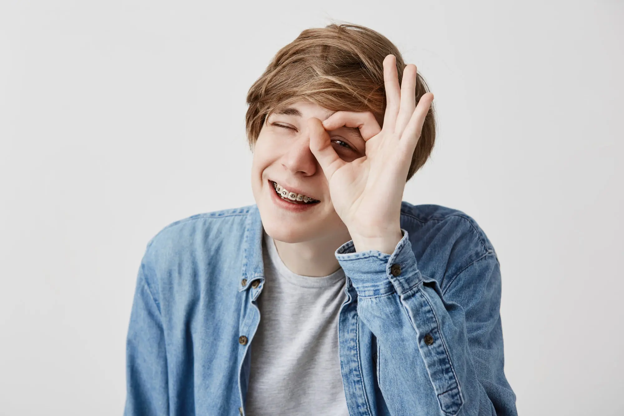 Smiling teen boy with affordable braces at Emerald City Orthodontics in Kirkland WA, makes an "OK" gesture around his eye.