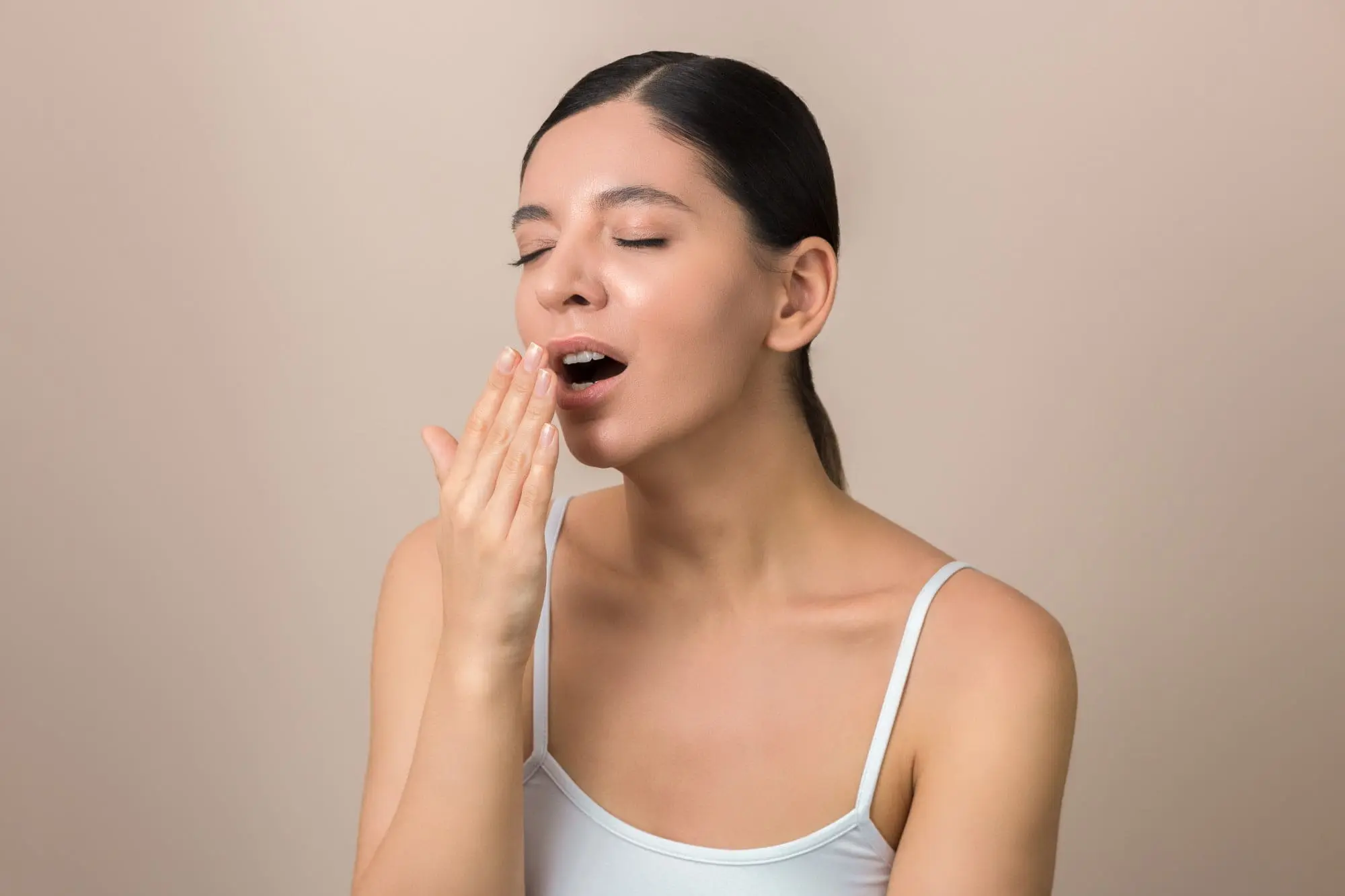 A woman in a white tank top with a slight underbite yawns, eyes closed, hand to mouth; Emerald City Orthodontics in Kirkland, WA.