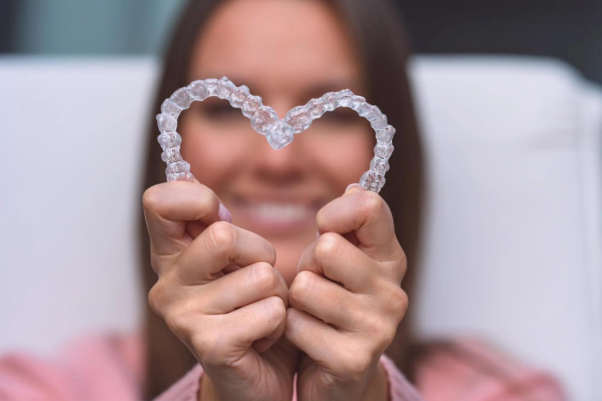 In Kirkland, WA, at Emerald City Orthodontics, a person forms a heart with two clear Invisalign aligners; face is blurred behind.
