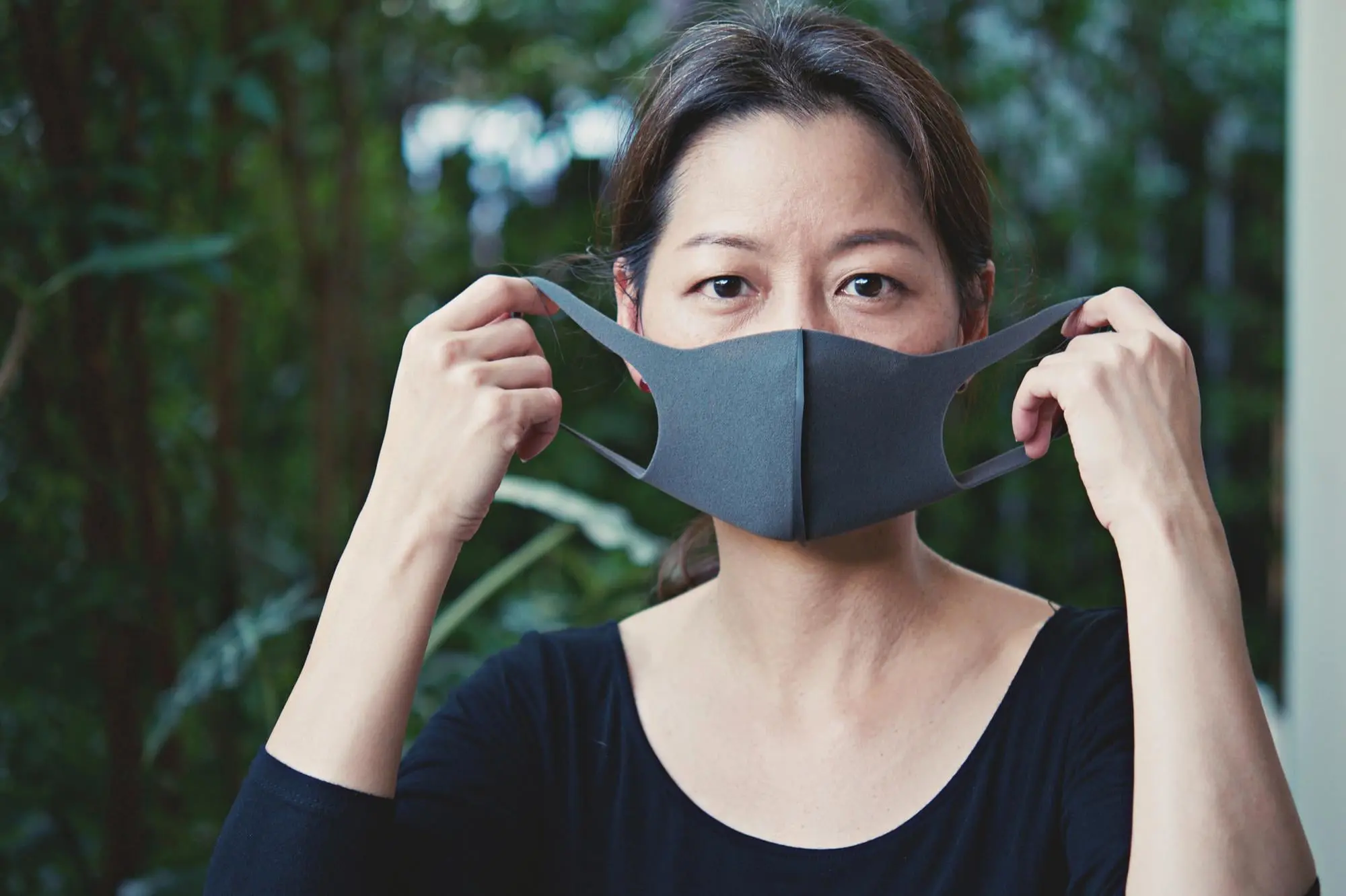 Outdoors in front of green foliage, a woman adjusts her black mask; possible mouth breather at Emerald City Orthodontics in Kirkland, WA.