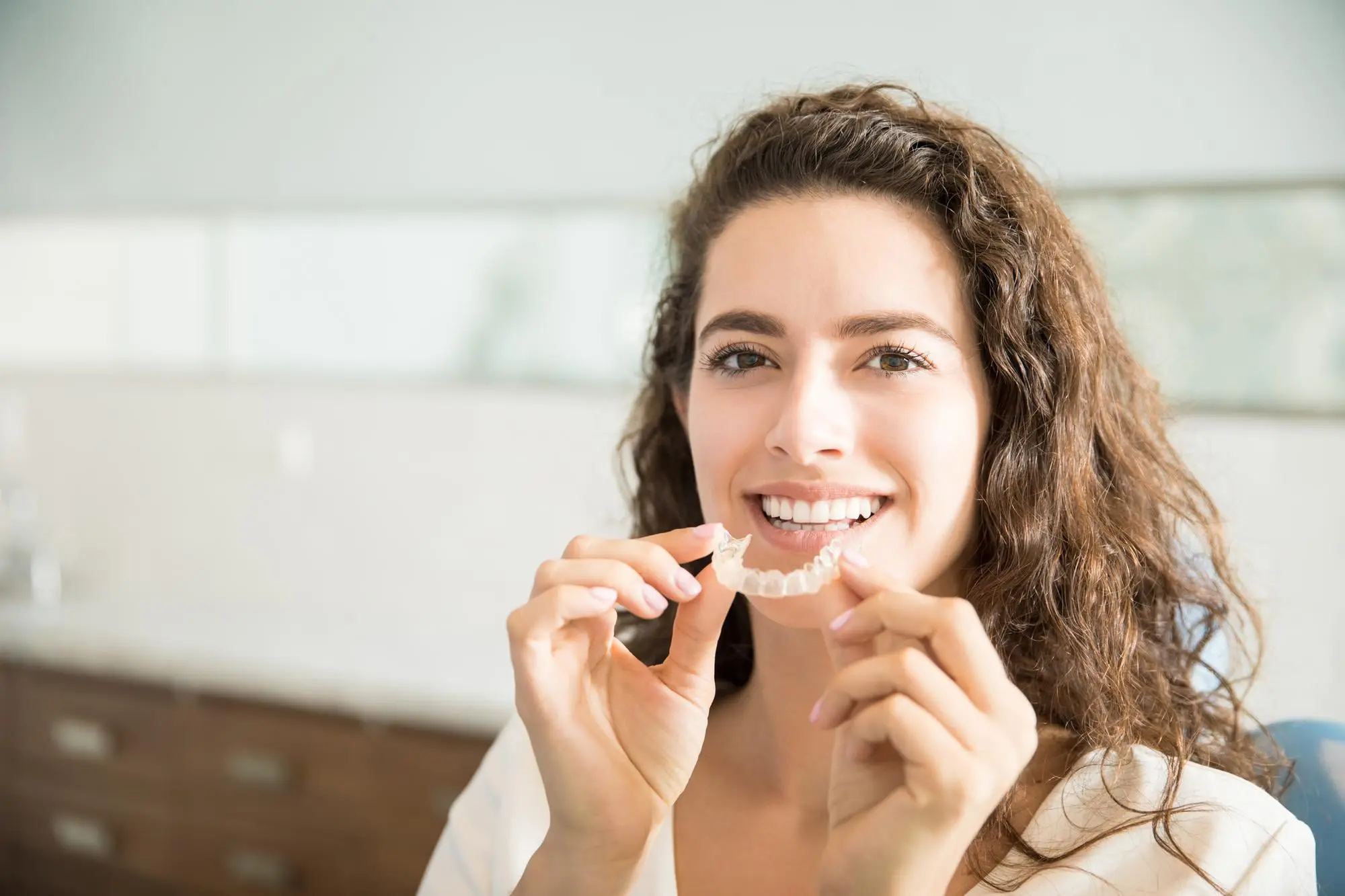 Smiling woman with curly hair holds an affordable Invisalign aligner at Emerald City Orthodontics in Kirkland, WA, indoors.