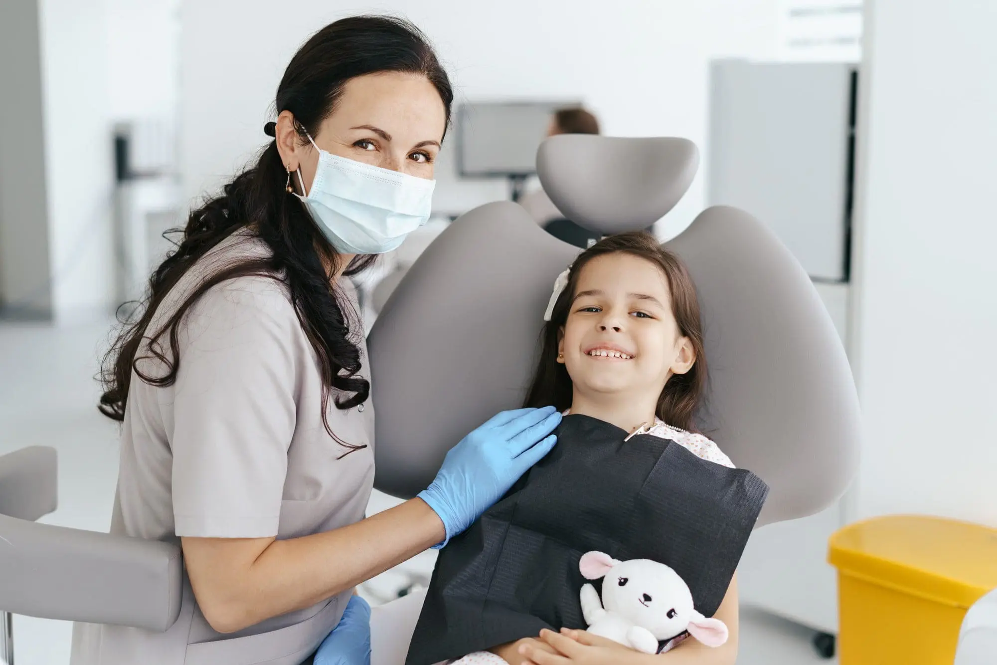A masked children's orthodontist at Emerald City Orthodontics in Kirkland, WA sits by a smiling girl holding a stuffed animal.