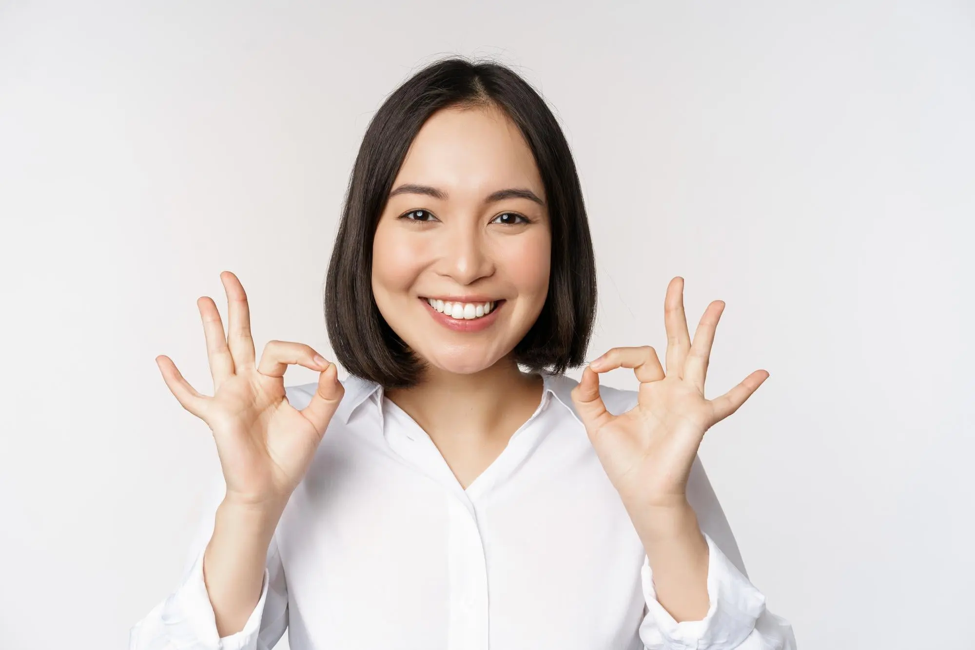 A woman with shoulder-length dark hair smiles and makes two "OK" signs, highlighting her Invisalign smile for Emerald City Orthodontics in Kirkland, WA.