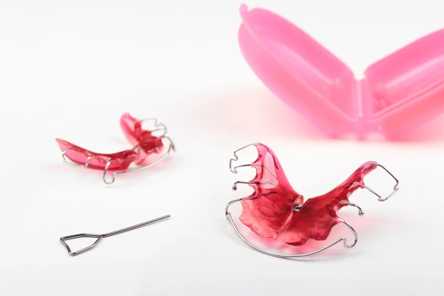 Displayed on a white surface are two red retainers, a metal key, and pink case used at Emerald City Orthodontics in Kirkland, WA.