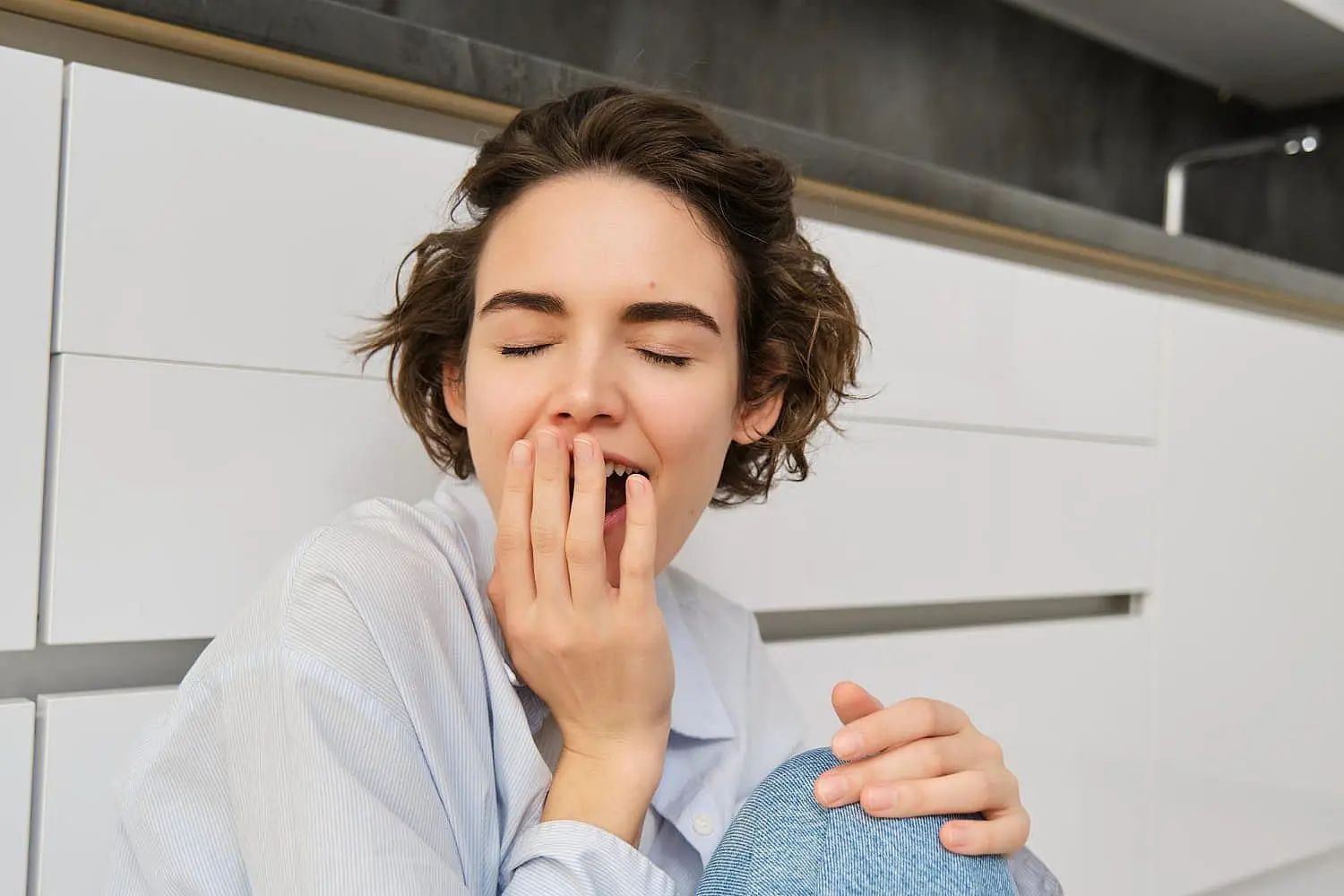 Seated on the floor in front of white cabinets, a short-haired person yawns, represent causes of mouth breathing at Emerald City Orthodontics in Kirkland, WA.