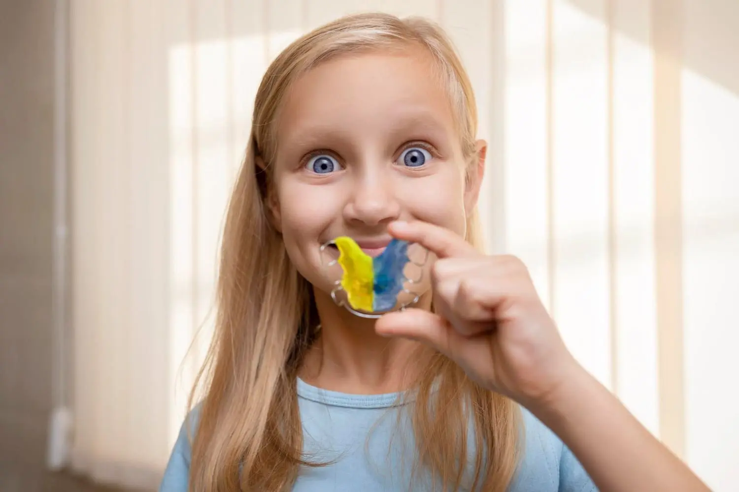 Smiling, a young girl holds a yellow and blue retainer after her Emerald City Orthodontics visit in Kirkland, WA.