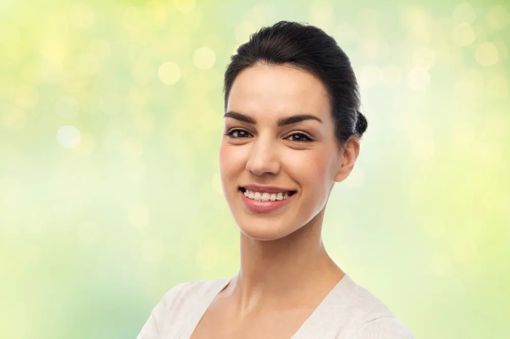 Smiling young woman with dark hair and clear braces at Emerald City Orthodontics in Kirkland, WA; blurred green background.