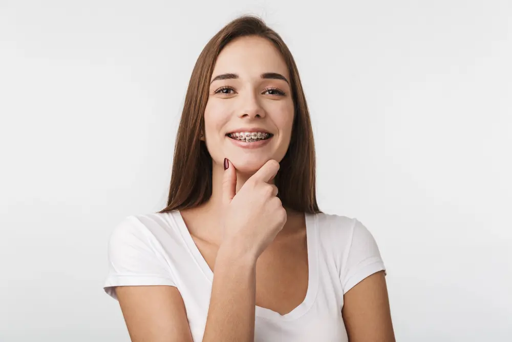 Smiling young woman with adult braces at Emerald City Orthodontics in Kirkland WA, touching her chin against a white background.