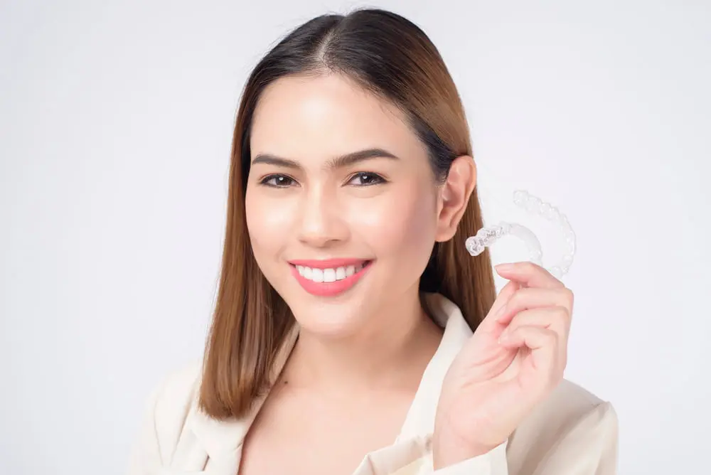 Smiling woman with straight brown hair holds Invisalign aligners at Emerald City Orthodontics in Kirkland WA, on white background.