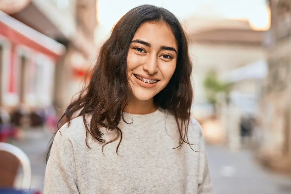 Smiling with metal braces, a young person stands on a city street near Emerald City Orthodontics in Kirkland, WA.