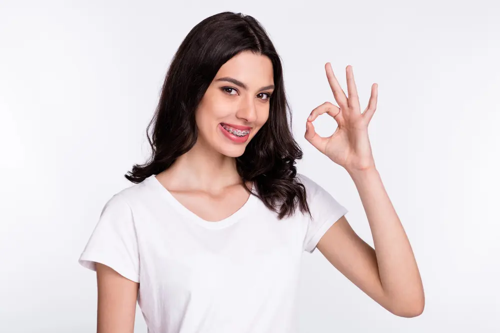 Smiling young woman with long brown hair and adult braces makes "OK" sign, promoting Emerald City Orthodontics in Kirkland, WA.