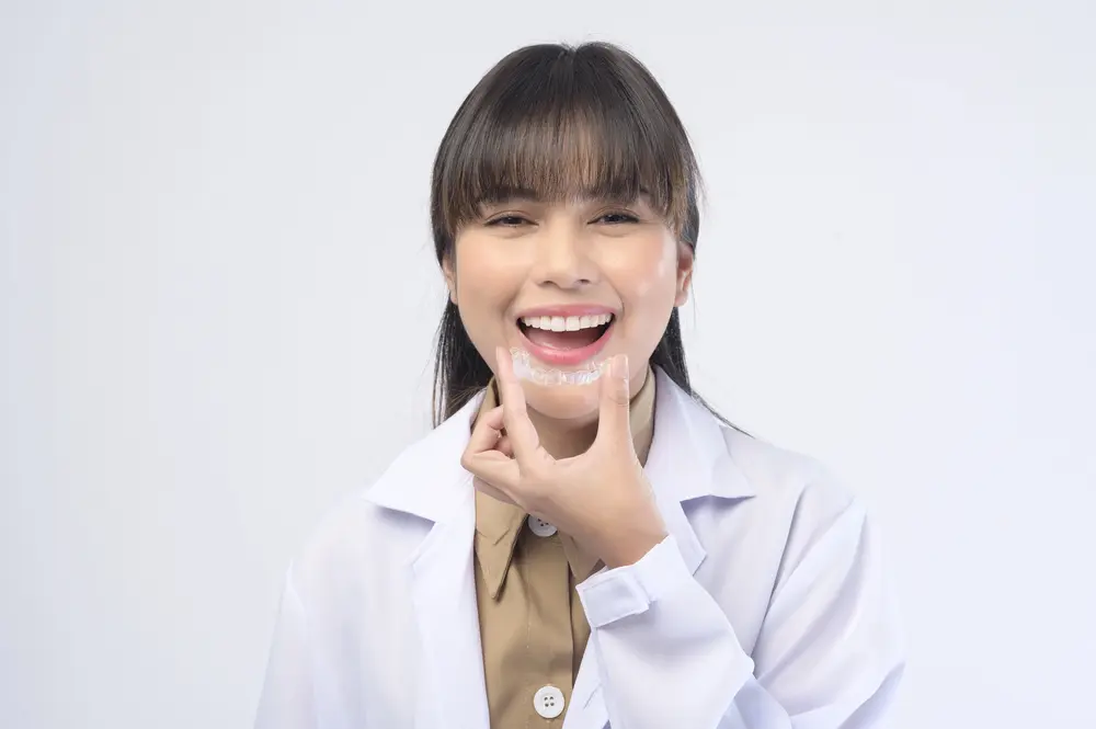 A woman in a lab coat smiles, applying cream to her chin and showcasing Invisalign aligners at Emerald City Orthodontics in Kirkland, WA.