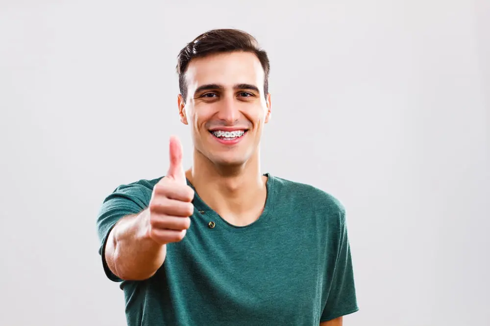 Smiling young man with metal braces in a green t-shirt gives a thumbs up, highlighting adult orthodontics at Emerald City Orthodontics in Kirkland, WA.