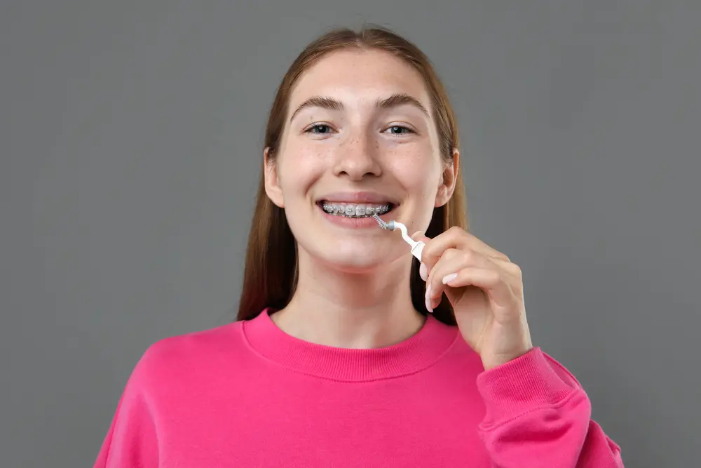 A young person with long hair and metal braces cleans their teeth with an interdental brush at Emerald City Orthodontics in Kirkland, WA.