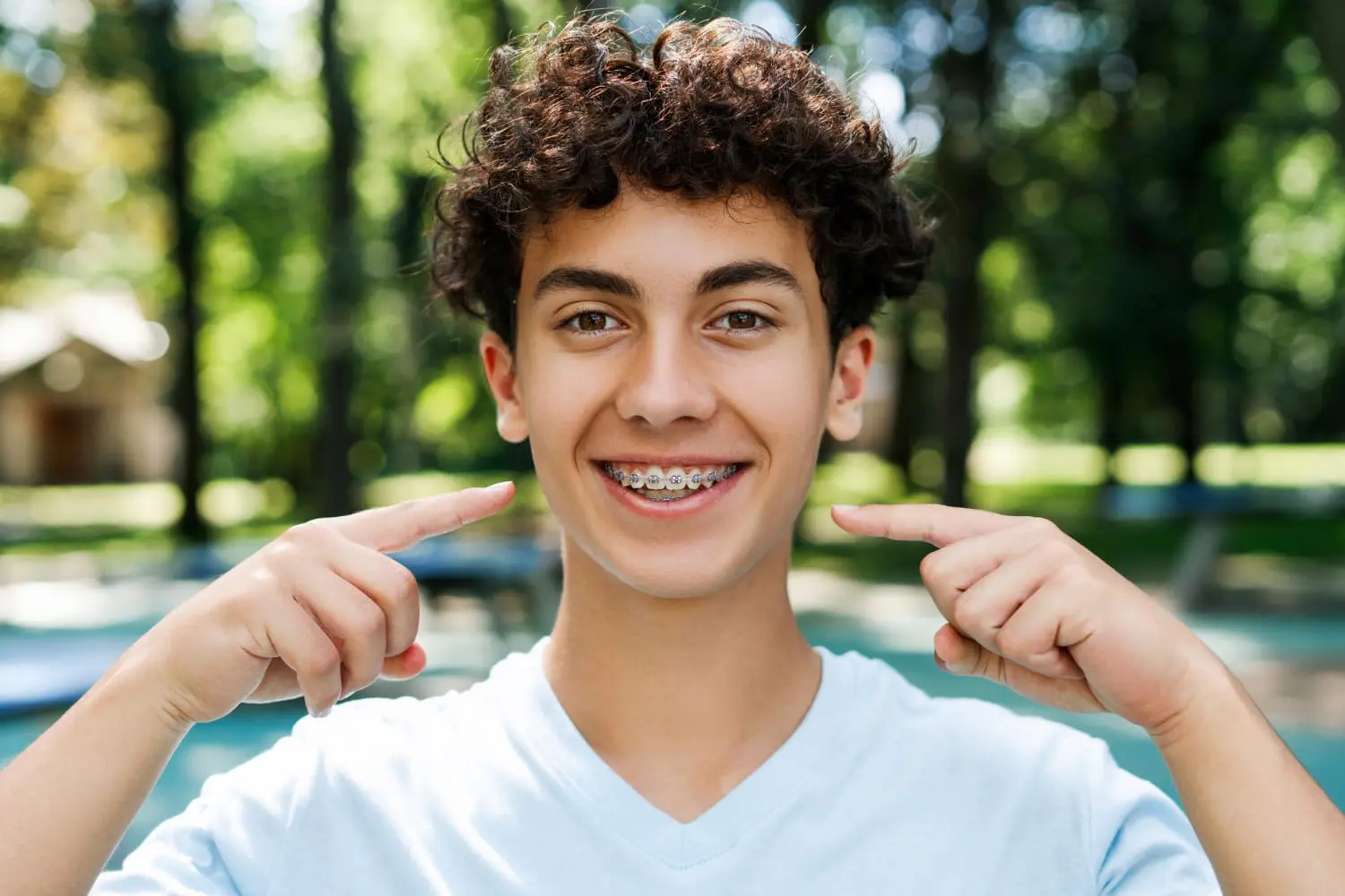 A teenage boy smiles and points to his metal braces outdoors for underbite treatment, promoting Emerald City Orthodontics in Kirkland, WA.