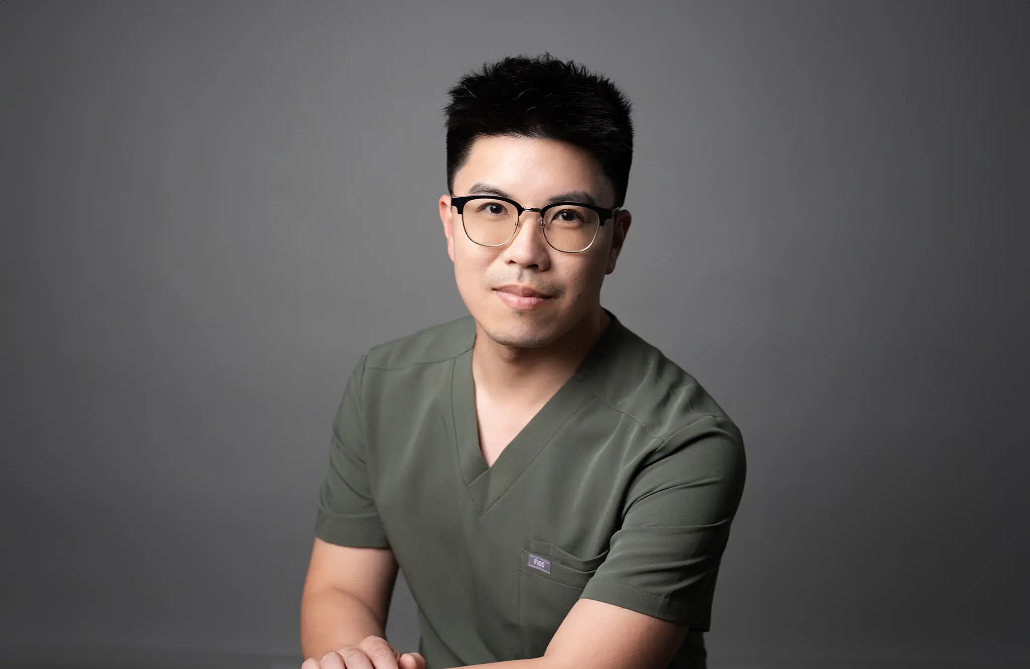 Dr. Hsin-Chuan (Dan) Pan, orthodontist expert at Emerald City Orthodontics in Kirkland WA, poses confidently in a professional portrait wearing green scrubs and glasses against a neutral gray background.