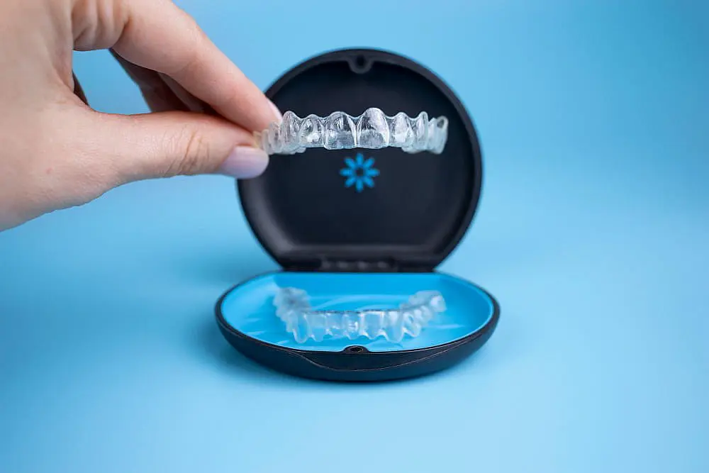 Showcasing Invisalign at Emerald City Orthodontics in Kirkland, WA: hand holds an aligner above an open case on a blue background.