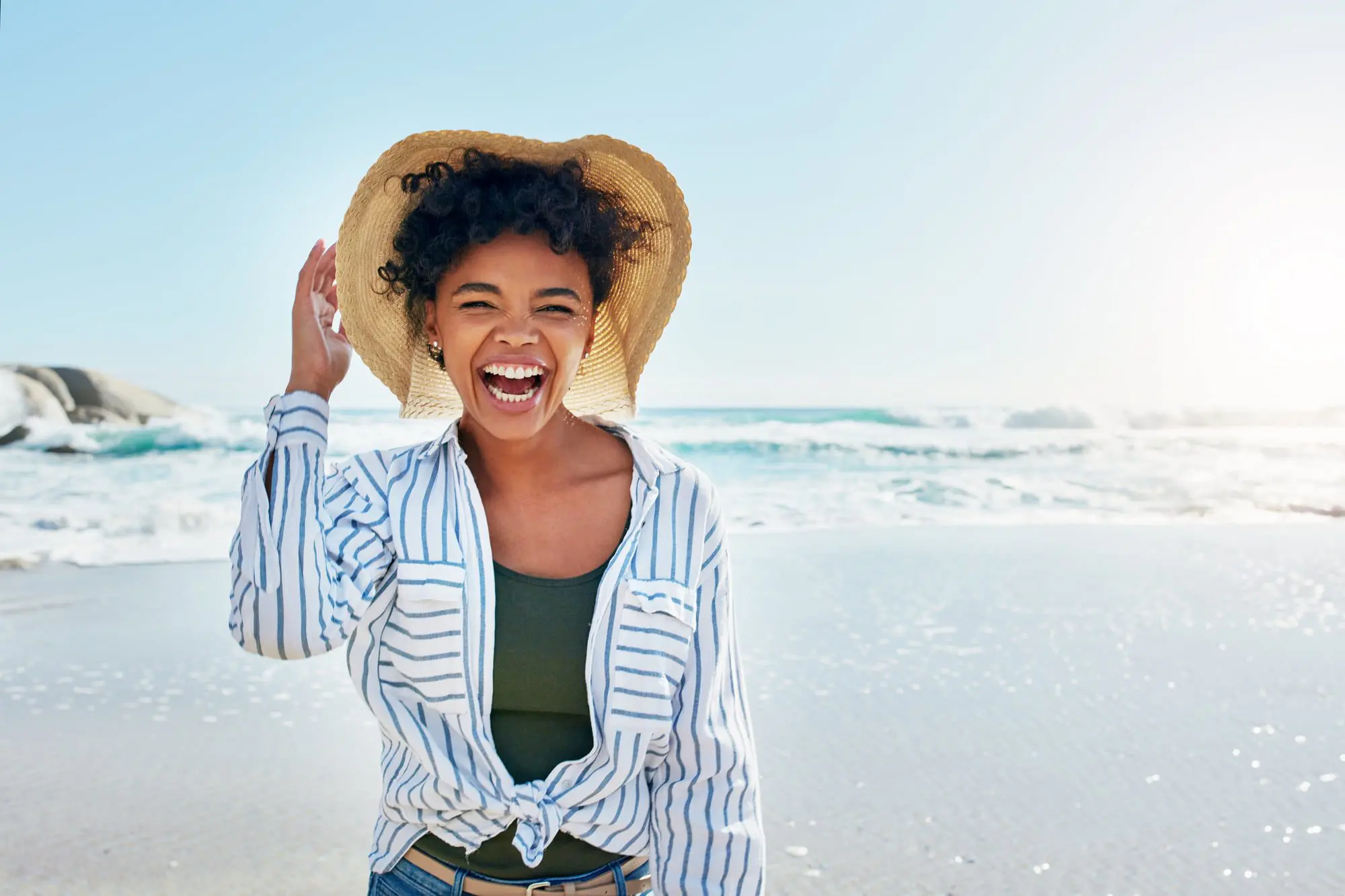 Emerald City Orthodontics Kirkland - Braces & Invisalign A smiling woman on a sunny beach, carefree in striped shirt and straw hat, thinks of the ocean, not braces cost at Emerald City Orthodontics in Kirkland, WA.