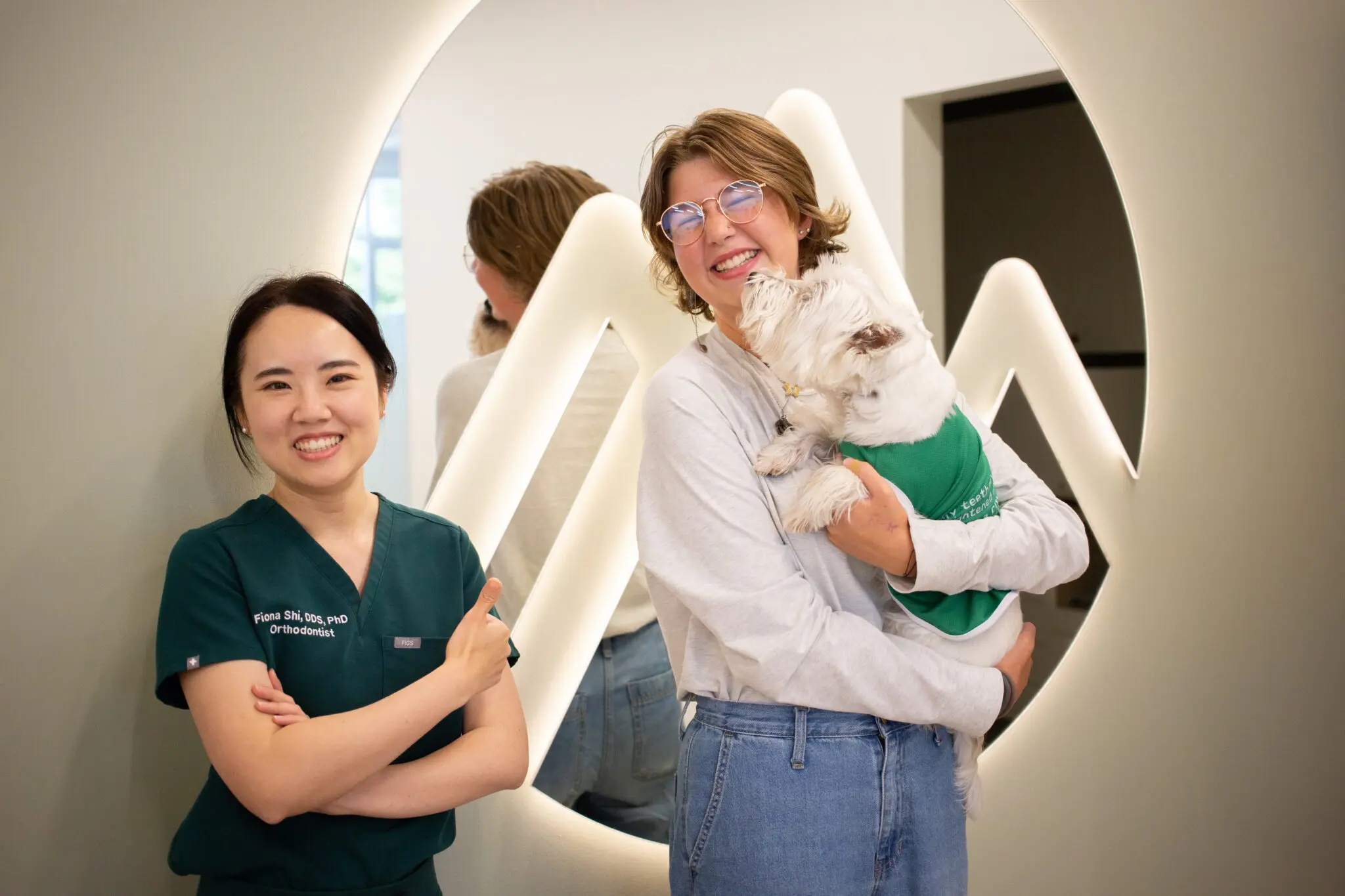 Two women, one in scrubs and one holding a white dog, smile by a modern wall at Emerald City Orthodontics in Kirkland, WA.