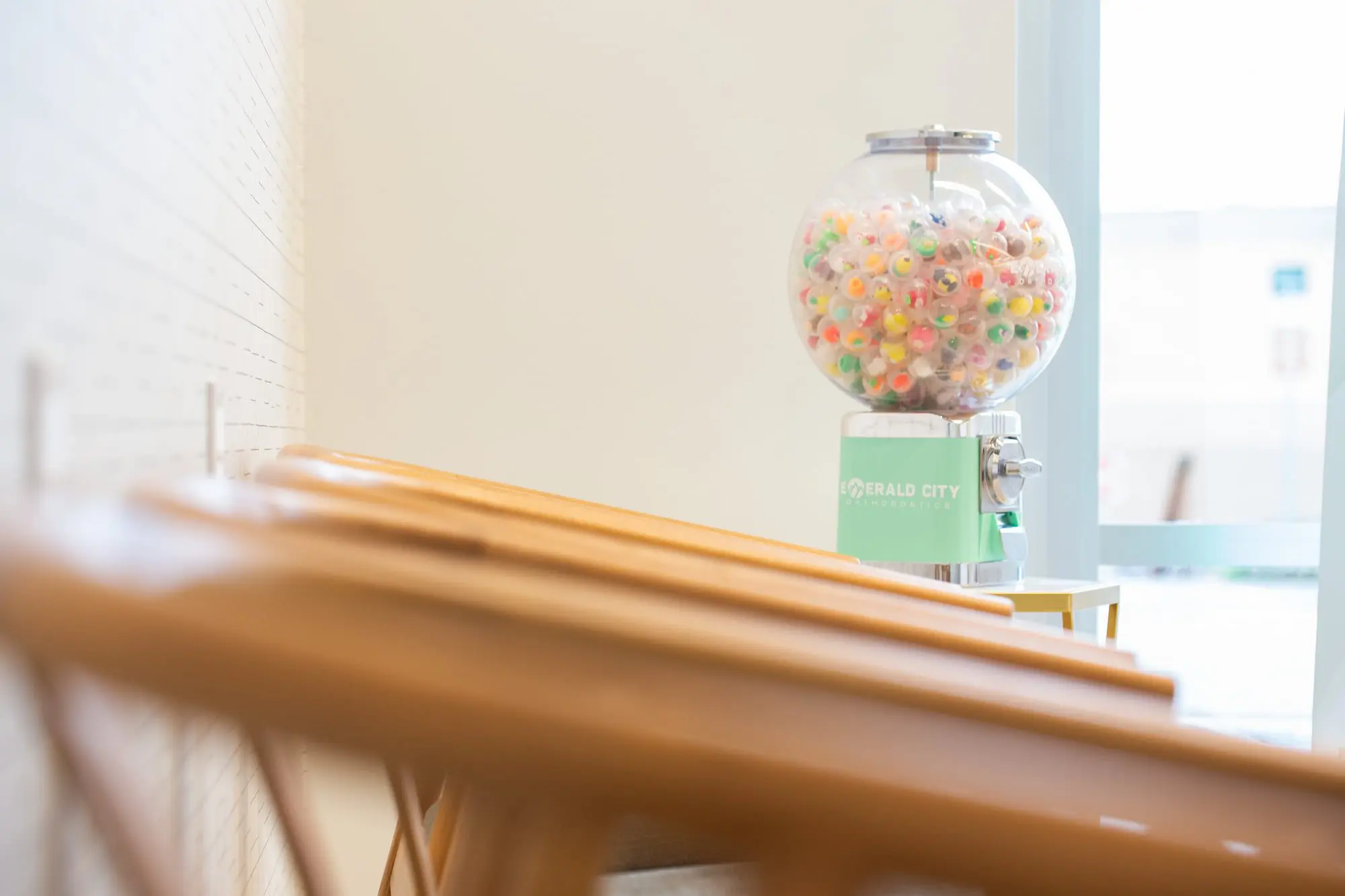 In Emerald City Orthodontics in Kirkland, WA, a colorful gumball machine sits by a window and empty wooden chairs in a bright clinic.