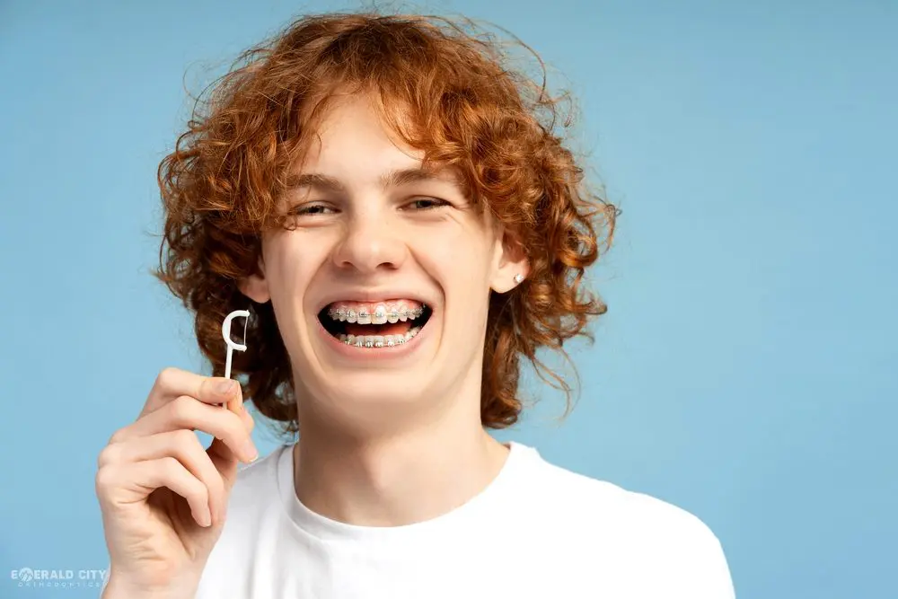 A smiling person with red curly hair and braces shows how to floss at Emerald City Orthodontics in Kirkland, WA, blue backdrop - How to Floss with Braces in Kirkland, WA