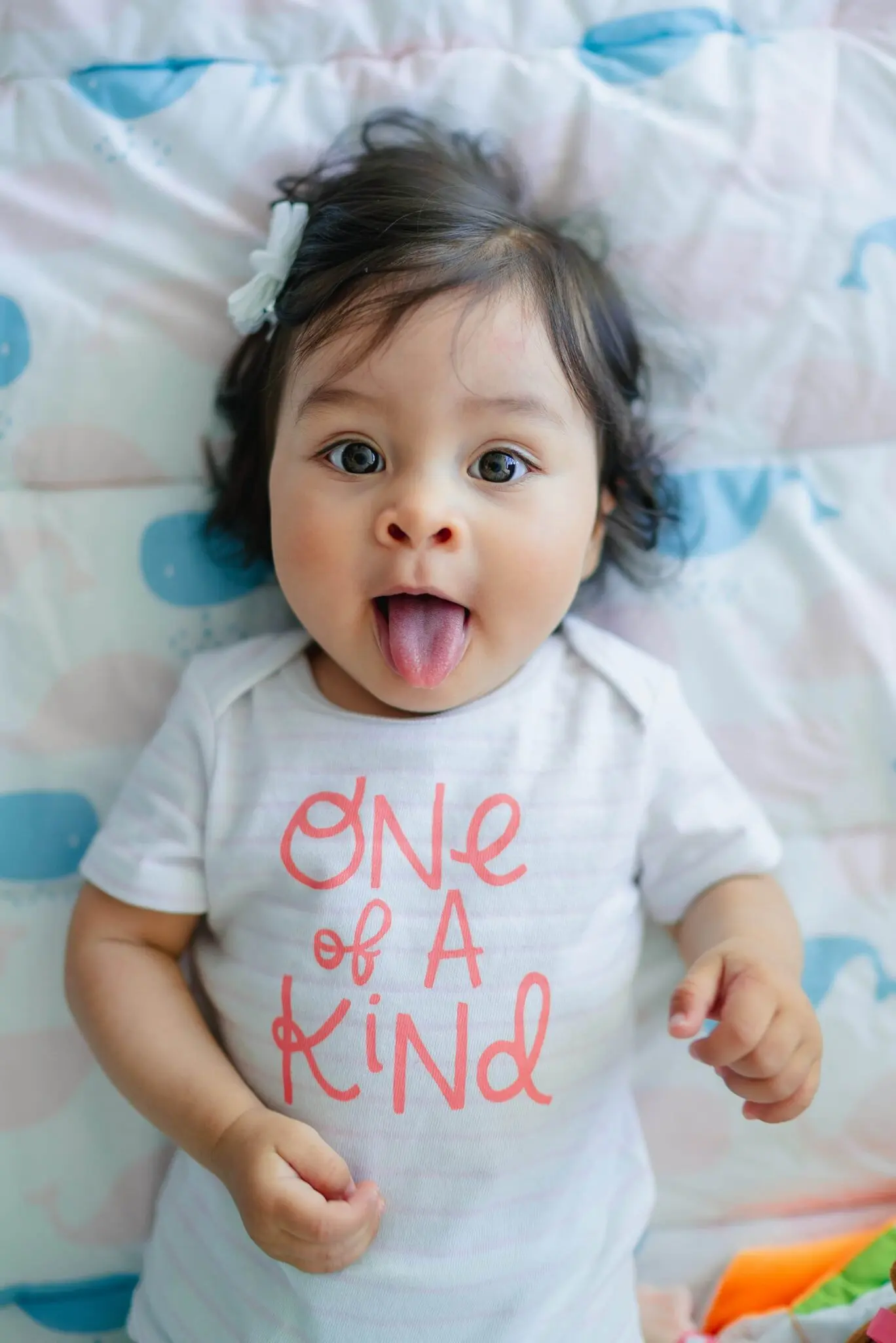 A baby with dark hair in a white bow and “One of a Kind” shirt shows a tongue thrust on a blanket at Emerald City Orthodontics in Kirkland, WA.