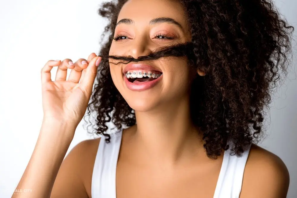 With a playful mustache pose, a smiling person with braces and curly hair showcases their smile at Emerald City Orthodontics in Kirkland, WA - How to Get Braces Glue Off Teeth in Kirkland, WA 