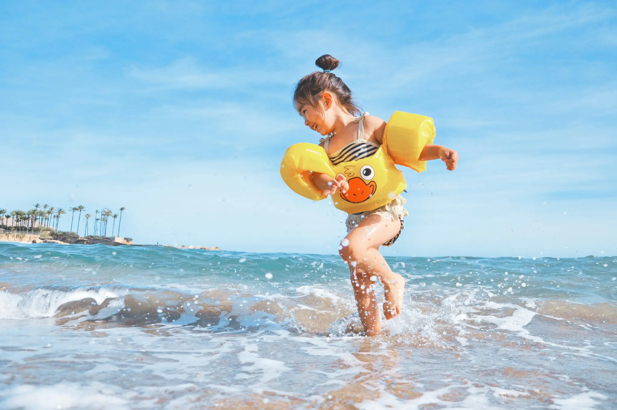 A young girl with yellow armbands plays in shallow waves, laughing as she practices tongue thrust for Emerald City Orthodontics in Kirkland, WA.