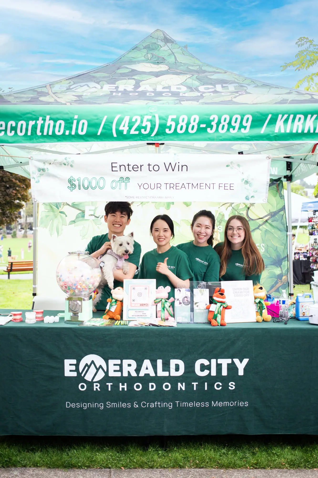 At an outdoor event in Kirkland, WA, four people and a dog stand by the Emerald City Orthodontics booth promoting careers and a $1000 giveaway.
