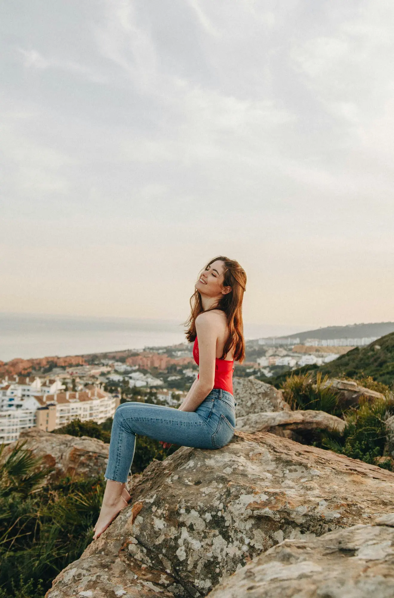Sitting on a large rock, a woman overlooks city and coastline views near greenery; Emerald City Orthodontics in Kirkland, WA.