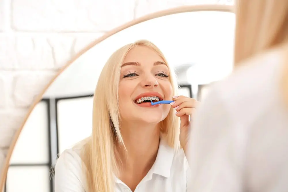 Brushing her teeth and flossing with braces in a mirror, a blonde woman follows Emerald City Orthodontics tips in Kirkland, WA - How to Floss with Braces in Kirkland, WA