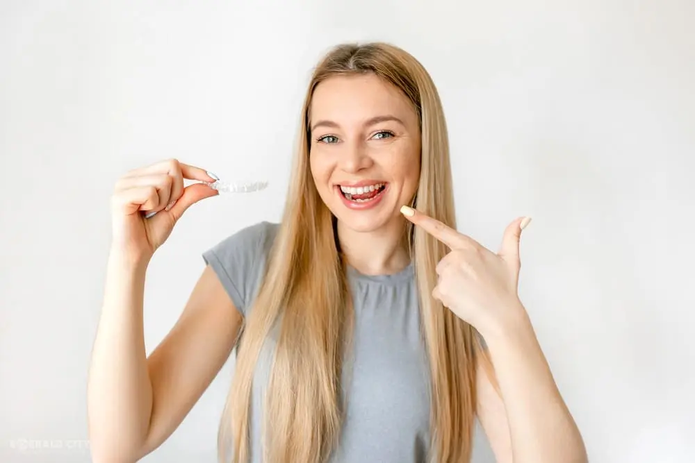 Smiling woman with long blonde hair points to her teeth and holds an Invisalign aligner at Emerald City Orthodontics in Kirkland, WA - Can Invisalign Fix Overbite in Kirkland, WA 