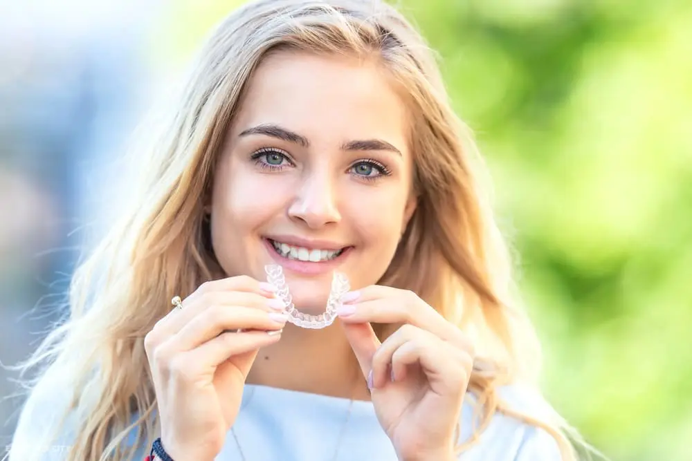 Smiling woman with long blonde hair holds an Invisalign aligner outdoors, promoting Emerald City Orthodontics in Kirkland, WA - How Does Invisalign Work in Kirkland, WA