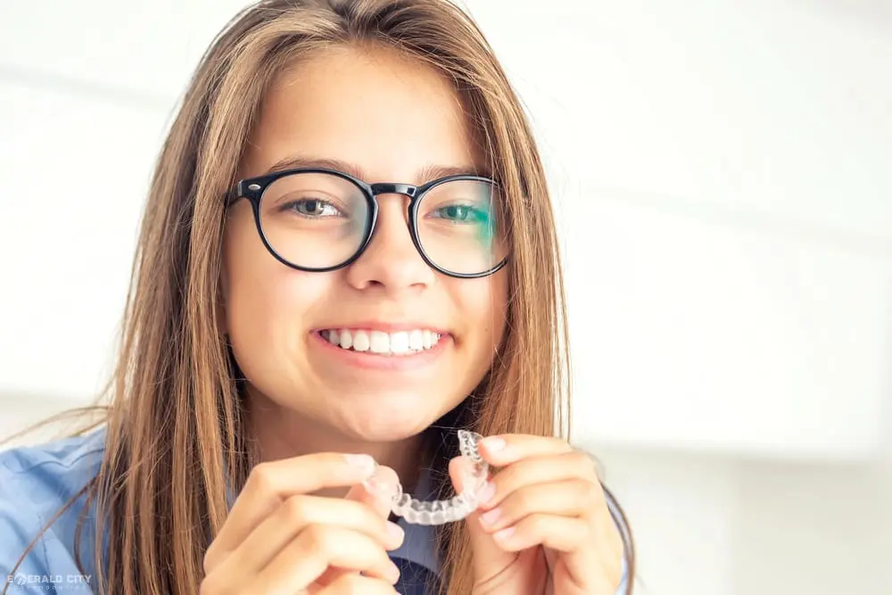 Young person with glasses holds a clear aligner, smiling wide at Emerald City Orthodontics in Kirkland, WA, showing Invisalign results - How Does Invisalign Work in Kirkland, WA
