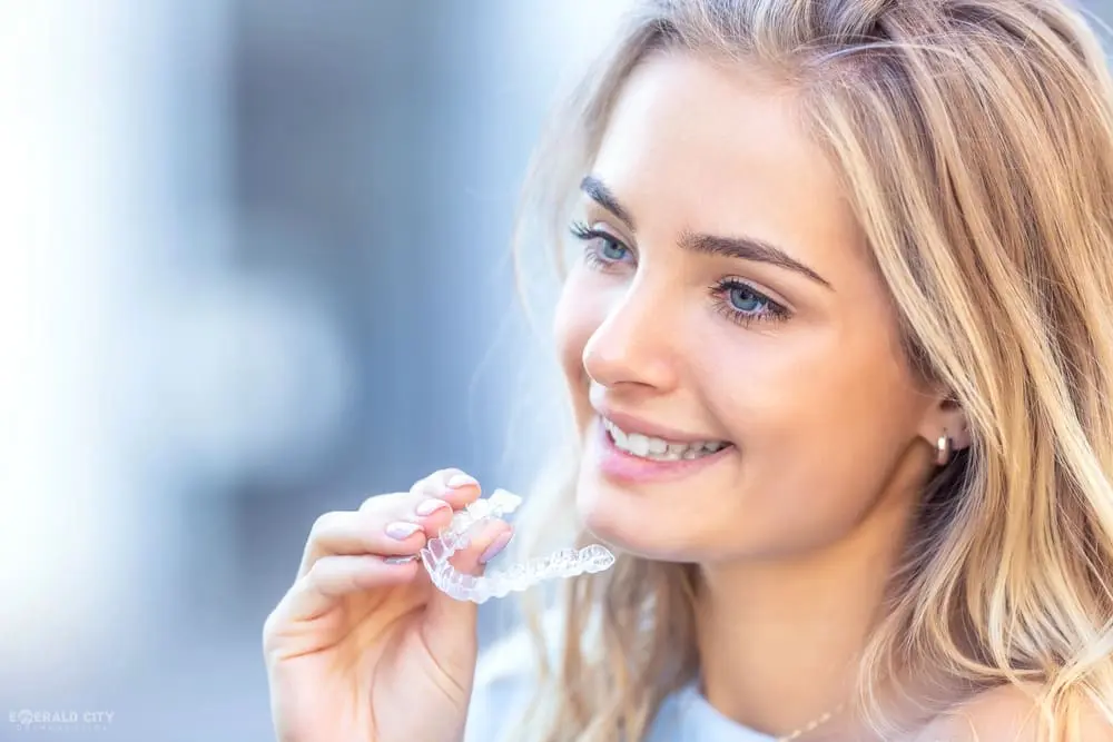 At Emerald City Orthodontics in Kirkland, WA, a smiling blonde woman holds a clear dental aligner near her mouth for overbite care - How to Fix Overbite in Kirkland, WA 