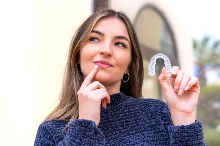 A woman outdoors in Kirkland, WA, holds a clear aligner and ponders overbite solutions at Emerald City Orthodontics - How to Fix Overbite in Kirkland, WA